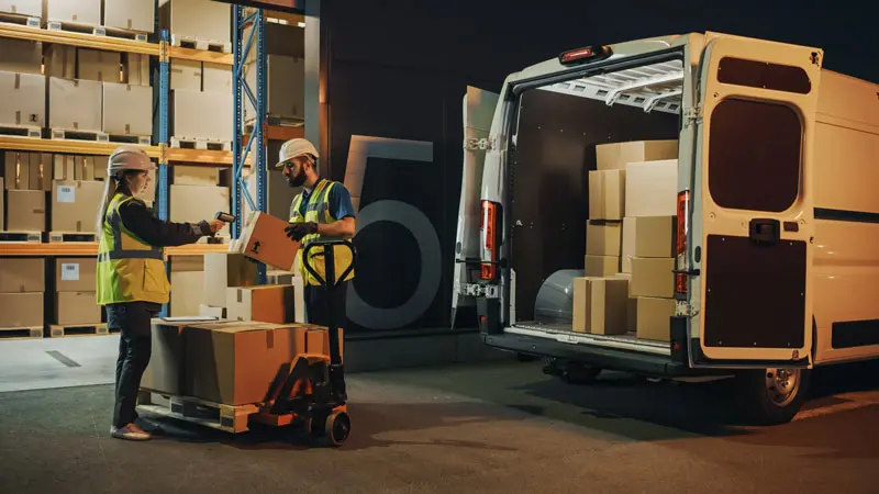 Trabajadores con equipo de seguridad cargando mercancía en una furgoneta en un centro logístico. La imagen representa la importancia de contar con una Póliza de Transporte de Mercancías para proteger los envíos ante pérdidas, daños o imprevistos durante el traslado