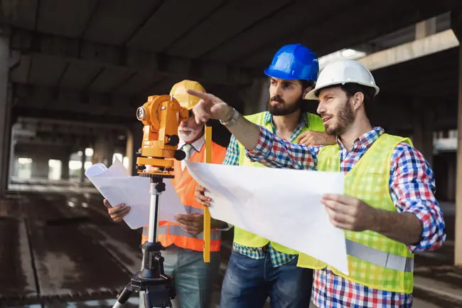 Ingenieros y supervisores de obra con cascos de seguridad revisando planos y utilizando un teodolito en una construcción. La imagen refleja la importancia de contar con una póliza Todo Riesgo Construcción y Montaje para proteger proyectos de infraestructura ante riesgos e imprevistos